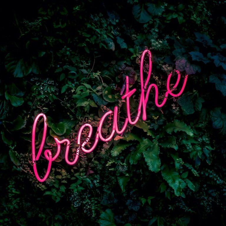 Neon pink "breathe" sign against a dark green leafy background.