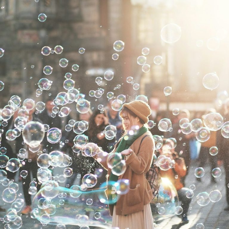 A woman in a hat stands among bubbles with a crowd in the background.